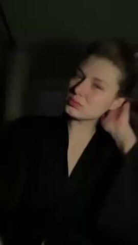 A woman with long, light brown hair pulled back into a ponytail and wearing a dark-colored robe poses for a video in a room illuminated by changing colored lights. The lighting shifts between blue, purple, and dark, revealing her fair complexion and facial expressions. She pouts, smiles, and smirks while facing the camera. A curtain or door is visible in the background.
