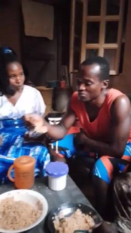The video shows four individuals gathered around a table, eating. Two people, a woman in a white blouse with a blue patterned skirt and a man in a red tank top and blue patterned shorts, are prominently featured, facing each other. They are holding up small objects, possibly phones or cameras. There is another person to the left of the woman, also holding a phone, and one more on the right of the man, whose body is partially visible. The table is covered with a blue and white checkered cloth. On the table, there are two plates filled with food, an orange mug, and a white container with a blue lid. The room appears to be dimly lit, with a window in the background. The walls are dark, and there are some miscellaneous items, like a towel hanging on a wall, that are visible in the background.