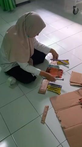 A person wearing a hijab and a light-colored shirt is sitting on a tiled floor, arranging small, individually wrapped items in rows. The items appear to be candy sticks or similar confections in various colors, including yellow, purple, orange, and green. The person is using cardboard pieces and tape, suggesting they are preparing to package or organize the items. There are several pieces of unfolded cardboard scattered around, as well as a roll of clear tape. The environment appears to be indoors, with a slightly green wall visible in the background and a medical cart with wheels on the right. The person looks at the camera and makes a peace sign.