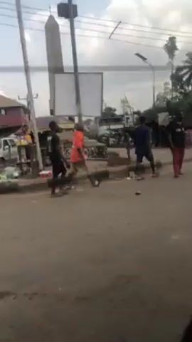 The video depicts an outdoor scene in what appears to be a busy street in a developing country. Several people are walking around, some wearing casual clothes and others wearing school uniforms. A yellow and black van drives by. There are simple shops or buildings visible along the side of the road, with corrugated iron roofs and wooden structures. A water tank can be seen atop one of the buildings. The road surface is unpaved, and there is litter scattered around. Palm trees and utility poles are visible in the background, with a partly cloudy sky above.