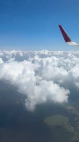 This media file captures a view from an airplane window as it flies above a blanket of white, fluffy clouds. The bright blue sky above is clear, suggesting a sunny day. Below the clouds, glimpses of the Earth's surface are visible, showing a patchwork of green fields and some scattered houses, hinting at a rural or suburban landscape. The wing of the airplane, with its distinctive red tip, is prominently featured in the upper right corner of the frame, anchoring the perspective. The sunlight illuminates the clouds from above, creating a sense of depth and texture. The overall impression is one of serene travel, with the vast expanse of the sky and clouds evoking a feeling of freedom and distance from the ground. There is no visible text in the media file. The location context of Parfenovo, Russia, suggests this aerial view is over that region. The time of day appears to be daytime, given the bright sunlight.