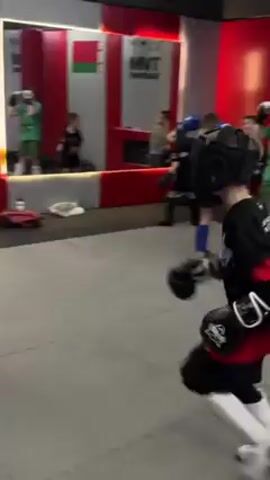 In a brightly lit training facility, two young boys wearing sparring gear engage in a kickboxing exercise. The boy closer to the camera is equipped with a full headgear, black gloves, shin guards, and a black and red shirt with an image of a fighter on the back. His opponent is similarly attired, albeit without shin guards. The sparring is dynamic, with each boy alternating between offensive kicks and defensive maneuvers. Other individuals are visible in the background, some engaging with punching bags. The room features gray mats, a sound system, and a wall decorated with red and white stripes. A mirror and the flag of Belarus are also visible.