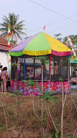 The video shows a colorful, octagonal inflatable attraction with a multi-colored roof. The sides are enclosed with black netting, and the base is decorated with a pink and blue pattern featuring spider-like figures. Inside, several children are visible, some appearing to be jumping or playing. Two adult women are standing outside the attraction, near the entrance. One woman, wearing a beige headscarf and a beige outfit, stands to the left of the entrance. To the right of the attraction, a person in a brown outfit is partially visible. The attraction is set up outdoors in what appears to be a rural area, with dry grass, sparse vegetation, and cassava plants in the foreground. A palm tree and buildings are visible in the background, along with Indonesian flags strung up, suggesting a festive occasion. The sky is overcast.