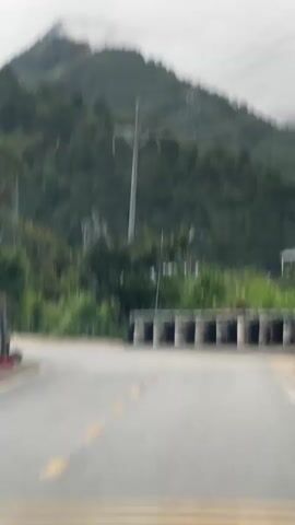 The video begins with a low-angle view of a mountain, partially obscured by clouds, with power lines crisscrossing the sky. The camera pans down to reveal a paved road leading towards the mountain, bordered by lush greenery and a makeshift barrier of tires. As the camera continues to pan, a yellow sign with Vietnamese and English text comes into focus, reading "Dốc dài nguy hiểm - Đi đúng làn / DANGEROUS LONG SLOPE - USE THE RIGHT LANE." A scooter and a person pushing a cart are seen moving down the road.