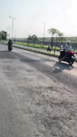 The video shows a paved road with several motorcycles. The road surface appears rough and under construction, with loose gravel in some areas. Three motorcycles are visible. The first is in the distance on the left, traveling away from the camera. The second is in the middle of the frame, also traveling away. The third motorcycle is only partially visible on the right side of the frame, also moving away. The environment appears to be outdoors with a clear sky and some trees and utility poles visible in the background.