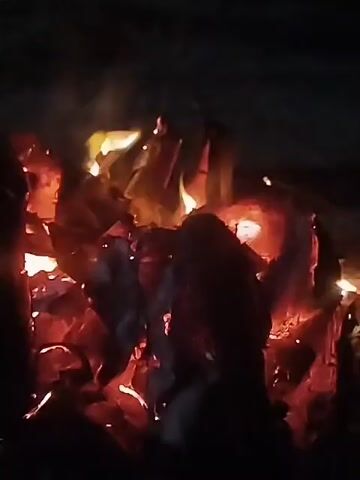 The video shows a close-up view of a burning fire in the dark. The flames are bright yellow and orange, and the embers glow red. Some smoke is rising from the fire. The camera angle is relatively steady, and the image appears to be captured indoors.
