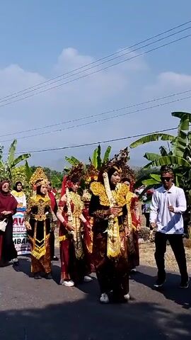 This is a vertical video of a procession on a paved road under a clear blue sky with some clouds. Several people are walking, some wearing traditional attire. The central figures are adorned in elaborate gold and red costumes, including headdresses that resemble crowns or elaborate hairpieces decorated with leaves or feathers. One individual in the center is wearing a gold necklace or garland. Some individuals are wearing hijabs. To the right, a man in a white shirt and black pants appears to be leading or gesturing. On the left, a banner is partially visible with text that includes "KARNA" and "MUT KE-63 MERDEKA." Banana trees and other greenery are visible on the sides of the road, and hills or mountains are in the background. The overall scene suggests a cultural event or parade.