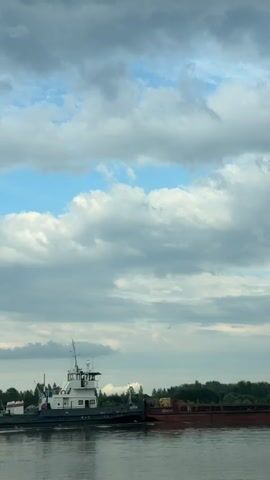 The video shows a tugboat pushing a barge on a body of water. The tugboat is white with a dark hull and has a tall cabin structure. The barge appears to be carrying some cargo, possibly wood or debris, and is mostly a dark red color. Trees and a distant shoreline are visible in the background. The sky is partly cloudy with patches of blue sky visible between the clouds. The water is calm, reflecting the sky and the boats. The scene appears to be set during the daytime, possibly late afternoon or early evening given the lighting. There is a Russian flag visible on the tugboat, and text is present on the side of the tugboat, though it is too blurry to read. The overall impression is of a working vessel engaged in transport on a river or similar waterway. The clouds suggest a dynamic weather system, but the overall mood is serene.