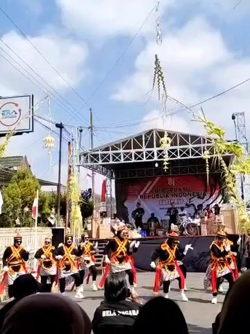 A group of dancers in traditional costumes perform in front of a stage decorated with Indonesian flags. The dancers are wearing black tops, red and white bottoms, and elaborate headpieces. They are moving in unison, with some of them holding their arms out to the sides. Behind them, a band is visible on the stage, and a banner that reads "81 DIRGAHAYU REPUBLIK INDONESIA" is displayed. The scene appears to be an outdoor celebration, possibly a parade or festival, with a crowd of spectators in the foreground. The weather is sunny, and the sky is clear.