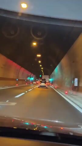 The video is a first-person view from inside a vehicle traveling through a tunnel. The tunnel is illuminated by overhead lights and lights embedded in the walls. There are other vehicles visible ahead, including a white car and a larger truck. The road has white lane markings and orange and green indicator lights along the sides. The interior of the vehicle, including the dashboard and windshield, is visible in the foreground. The scene appears to be at night or in low light conditions.