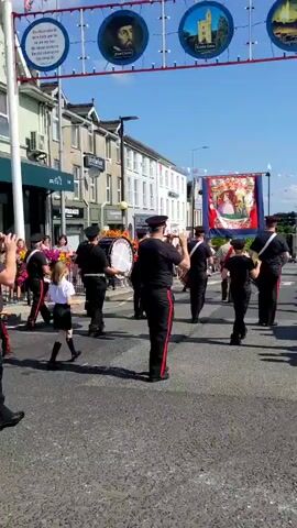 The video shows a parade on a street under a bright sky. A group of people in uniform are marching and playing instruments, including drums and what appear to be flutes. They are wearing black uniforms with red stripes on their pants and hats. Some of the marchers are carrying banners. There are spectators on the side of the street. The street is paved, and there are buildings on either side. The time stamp indicates the video is very short, just a few seconds long.