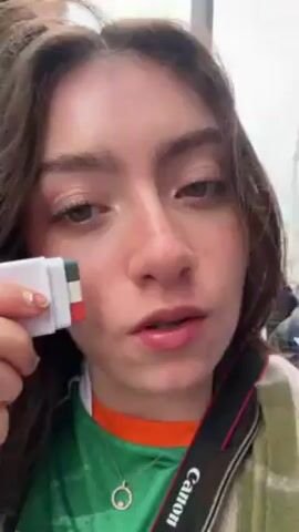 A young woman applies face paint in the colors of the Irish flag to her cheek with a small, multi-colored crayon stick. She is outdoors, wearing a green jersey with an orange collar and a necklace with a circular pendant. She has brown hair, fair skin, and a nose ring. A camera is visible on a strap across her chest. In the background, people are walking on a city street. She makes a kissing face and poses.