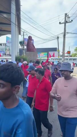The video shows a political demonstration in what appears to be a city street.  The demonstrators, a diverse group of adults and children, are primarily dressed in red clothing, with many wearing red hats or carrying red flags. The marchers are walking on a sidewalk and a street, with buildings lining the route. Some buildings have signs in a language other than English. A statue of a figure is visible in the distance. A man in a bucket hat is prominently featured in the foreground, looking down. Other individuals hold umbrellas and cell phones.
