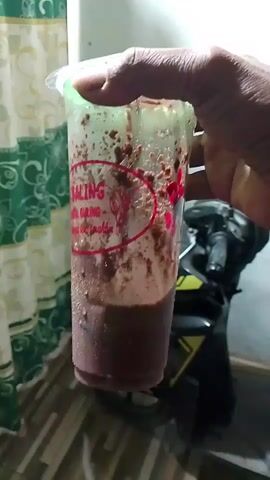 A hand is holding a clear plastic cup filled with a dark, frothy beverage. The cup has red writing on it. The hand is shaking the cup vigorously, causing the contents to swirl and splash against the sides. The background is blurred but appears to be indoors with a patterned curtain on the left and a motorcycle partly visible on the right. The lighting is dim.