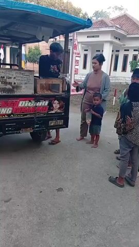 A young man stands behind a food cart with a blue awning. He is interacting with a woman and a young boy who are standing in front of the cart. The woman appears to be handing something to the boy, who is holding a plastic bag. Several other children are visible in the background. The food cart is decorated with colorful lettering and an image of a woman. The scene appears to be taking place outdoors, possibly on a street or in a public area, with buildings and trees in the background.