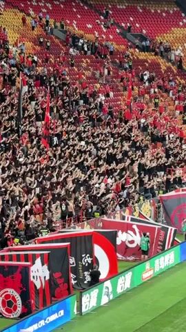 The video shows a large crowd of sports fans in a stadium, predominantly wearing black shirts. Most of the fans are standing and raising both arms in unison, creating a synchronized movement. In the foreground, there are banners with black, red, and white colors. A security guard is visible, wearing a green jacket, standing near the banners. Some individuals are seen wearing sports jerseys. The stadium's seats are filled with a mix of red and yellow colors. The atmosphere appears energetic and spirited, suggesting a sporting event with enthusiastic support from the fans.