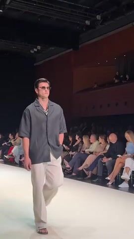 A male model walks down a runway wearing a dark grey short-sleeved button-down shirt, light beige pants, and sandals. He has on sunglasses and a patterned scarf visible around his neck. The runway is white, and the background features an audience seated in tiered seating, with wooden accents and dark ceilings. The lighting is professional and directed towards the runway.