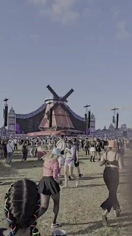 The video shows a wide shot of an outdoor festival with a large crowd gathered. In the background, a massive stage designed to look like a windmill is the focal point. The windmill stage has large video screens on its facade, displaying vibrant purple lights and graphics. Flanking the windmill stage are tall speaker towers and smaller architectural elements that mimic a medieval village. The ground is covered in dry grass, and the sky is a clear blue with a few scattered clouds. In the foreground, two individuals are walking away from the camera, with their backs to the viewer. The person in the immediate foreground has dark hair styled in two long braids with pink accents, and is wearing a black shirt and a black bag slung across their body. The person to their right is wearing a black t-shirt with a graphic on the back and khaki pants. Numerous other festival-goers are visible in the middle ground, walking, standing, and interacting. The overall atmosphere appears to be that of a lively outdoor event under a bright, sunny sky.