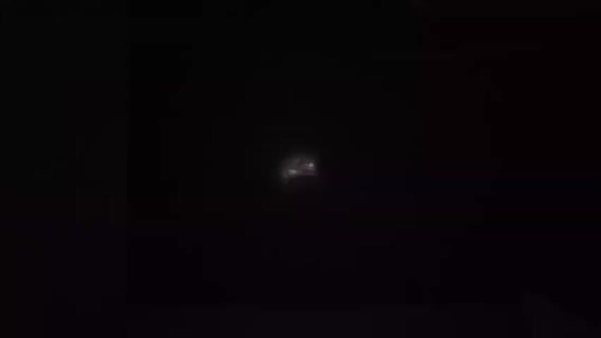 A hand holds and sprays a dark can in a dark environment. The spray is visible in the darkness.