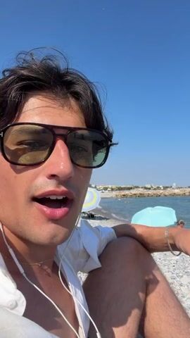This media file features a close-up of a man with dark, wavy hair and sunglasses, wearing a white shirt. He is sitting on a pebble beach on a sunny day. In the background, the blue sea stretches out to the horizon, with a few boats and swimmers visible. Palm trees and buildings can be seen further down the coastline. The man is speaking and gesturing, appearing relaxed and engaged. The overall mood is casual and summery, suggesting a vacation or leisure activity. The time of day is clearly daytime due to the bright sunlight and clear blue sky. The location is identified as Cagnes-sur-Mer, France, which is consistent with the presence of palm trees and Mediterranean architecture.