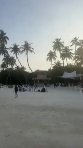 The video shows a picturesque beach scene. Initially, a wedding reception is visible, with tables and chairs set up on the sand. A person in dark clothing is walking towards the left side of the frame. Tall palm trees provide shade and add to the tropical ambiance. The scene then shifts to the right, revealing a long pier extending into the turquoise water. Overwater bungalows line the pier, stretching into the distance. The camera continues to pan to the right, and a small red boat comes into view, gently rocking in the calm water. The sky is a pale blue, suggesting either early morning or late afternoon.