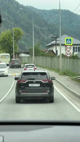 The video shows the view from the driver's seat of a car driving on a road. The car is following a black SUV. There are other cars on the road, including a white SUV and a white car. In the background, there are mountains with trees. There are also houses and a pedestrian crossing. The weather appears to be overcast.