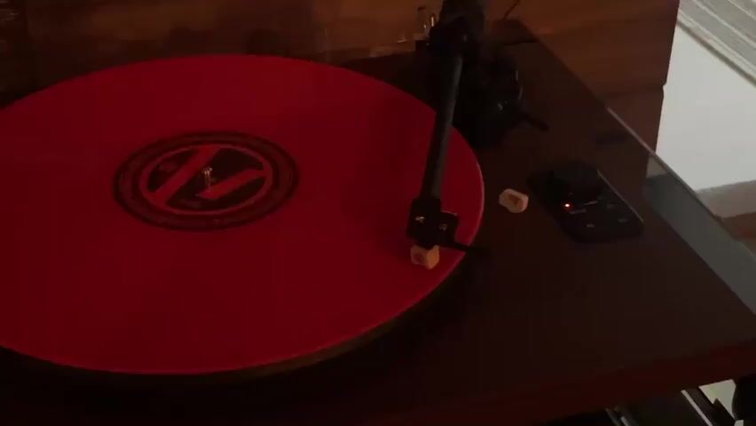 A red vinyl record is playing on a turntable. The turntable is dark brown and sits on a surface that appears to be a wooden table. To the right of the turntable, there is a small black remote control with a red light, and two white objects that look like ear buds or small pills. The background is dimly lit and appears to be a room with some wooden paneling and a white blinds. The video is shot from a slightly elevated angle, looking down at the turntable.