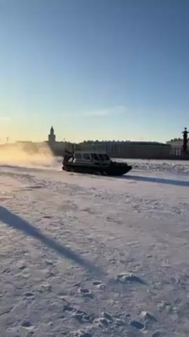 The video shows a black hovercraft driving across a snow-covered landscape. In the background, there are buildings visible under a clear sky. As the hovercraft moves, it kicks up a cloud of snow behind it. The hovercraft has writing on the side, partially visible. The light suggests it could be either early morning or late afternoon.