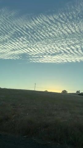 This is a video of a landscape filmed from a moving vehicle. The sky is blue with scattered white clouds forming a wavy pattern. A grassy hill slopes upwards from the foreground. There are a few trees on the hill, and telephone poles with power lines are visible. The sun is low on the horizon, casting a yellow glow. The foreground appears to be a road.