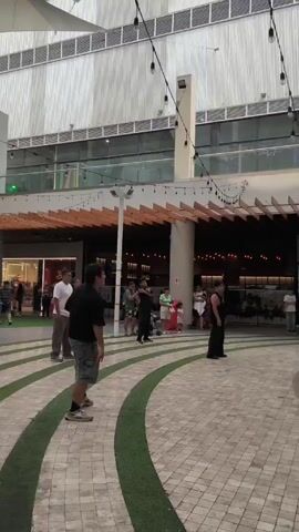The video shows a group of people seemingly practicing a dance in an outdoor plaza. There are about 8-10 individuals spaced apart, dressed in casual attire. They are performing synchronized arm movements. The plaza has a patterned brick floor with curved green inlays, and is surrounded by modern buildings with retail spaces. String lights hang above, adding to the atmosphere. Several other people are walking by or observing the dance practice. The weather appears to be sunny.