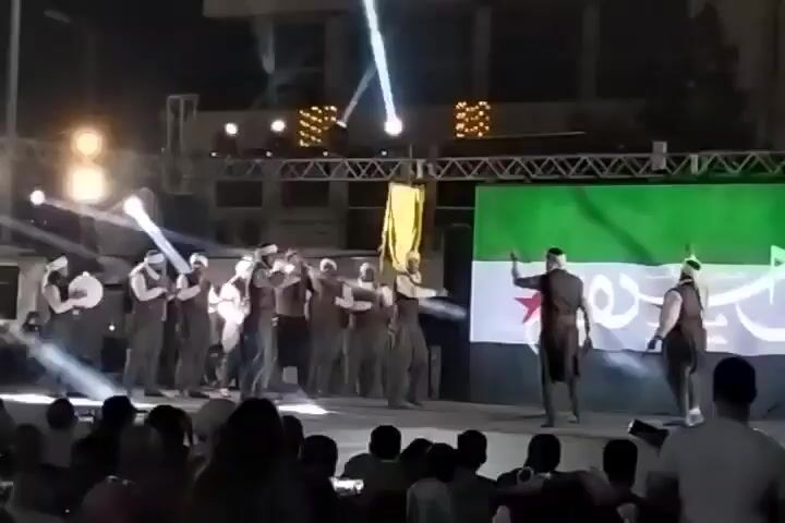 A group of men are performing on an outdoor stage. They are wearing dark vests and loose pants with white headbands. They are holding hands and moving in unison. Some of the men are playing drums. Behind them is a large screen displaying an image of a flag that is green, white, and black with a red star.  The stage is illuminated with bright white spotlights.  A crowd is gathered in front of the stage, watching the performance.