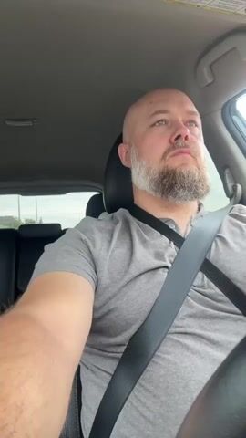 A man with a bald head and a light-colored beard is seated in the driver's seat of a car. He is wearing a grey t-shirt and a dark seatbelt across his chest. He looks forward and slightly upward, appearing to be driving. The interior of the car, including a headrest and part of the ceiling, is visible. Music is playing.