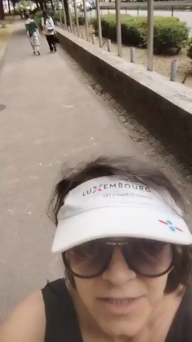This is a selfie-style video taken outdoors, likely on a paved walking path in a park or along a street in Vila Nova de Famalicão, Portugal. The person in the foreground is wearing sunglasses and a white visor with the text "LUXEMBOURG LET'S MAKE IT HAPPEN" printed on it, along with a stylized logo. They are smiling slightly at the camera.

In the background, a paved path stretches into the distance. Several other individuals are visible walking away from the camera along this path. The setting appears to be a park or a green area, with trees and bushes lining the path. There are also some metal posts and a low wall or railing along one side of the path. The ground beside the path is covered in dry leaves and dirt, suggesting it might be late spring or summer. The lighting indicates it is daytime, possibly late morning or afternoon, with overcast skies contributing to diffused lighting. The overall mood seems relaxed and pleasant, consistent with a walk in a park.
