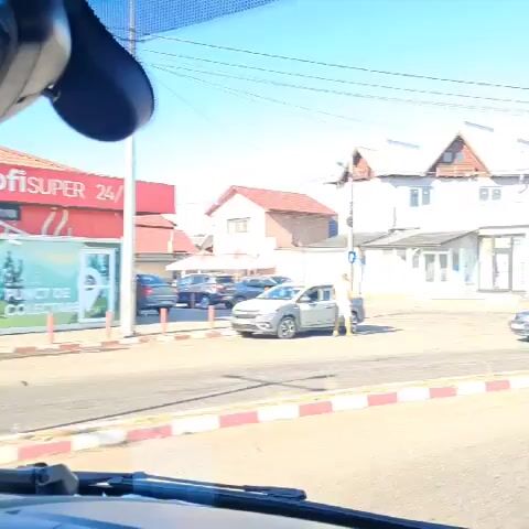 The video captures a street scene from the perspective of a vehicle. The road is paved, with white lane markings. A pedestrian, wearing a yellow shirt and dark shorts, stands in the middle of the intersection, gesturing towards a gray taxi. Another vehicle, a light blue sedan, passes by. Buildings line the street, some with multiple stories and balconies. A large white truck carrying construction materials is parked on the side of the road. Power lines crisscross the bright blue sky. The overall environment appears to be a town or city with commercial buildings and residential structures.