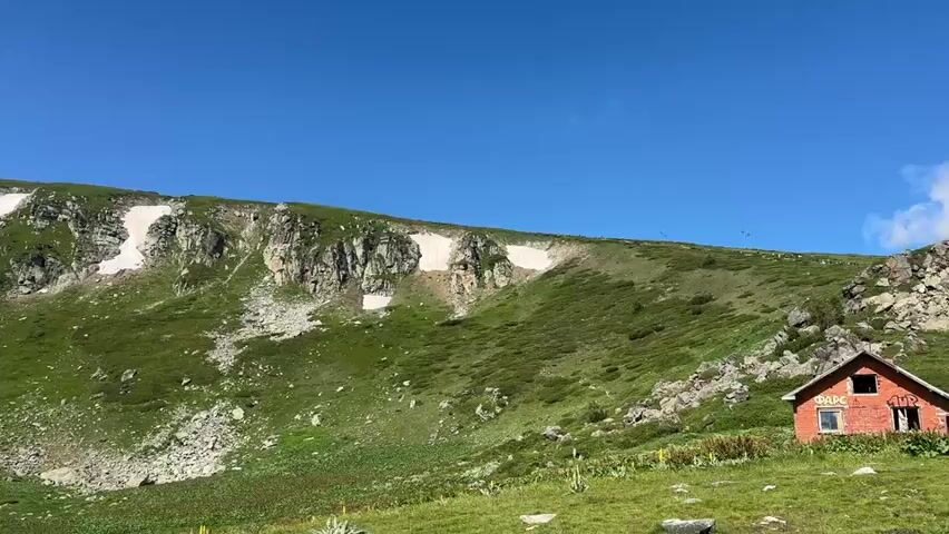 The video pans across a mountainous landscape under a clear blue sky with a few scattered clouds. The slopes are covered in lush green grass and low-lying shrubs. Patches of snow are visible on the steeper, rockier sections of the mountainside. In the foreground on the right, a small, dilapidated orange building with a dark roof stands. A path winds its way through the green terrain in the mid-ground. Further in the distance, more rugged mountain peaks are visible, some also capped with snow. The overall impression is a remote, natural environment in good weather conditions.