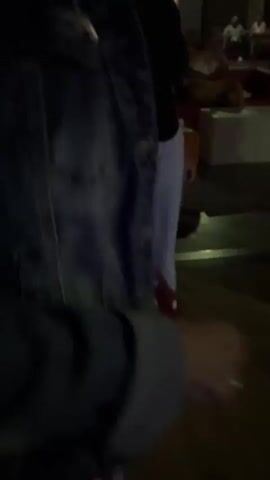 The video shows a dimly lit indoor space, possibly a club or event venue, with several people visible. The camera is in motion, suggesting it is being held by someone walking or moving around. The lighting is dynamic, with some areas illuminated by red and purple lights, and a large screen displaying a colorful, possibly abstract or music-related visual. People are seen in various states of activity, some standing and talking, others appearing to be dancing or observing the screen. The overall atmosphere appears to be social and possibly festive, with a focus on entertainment and gathering.