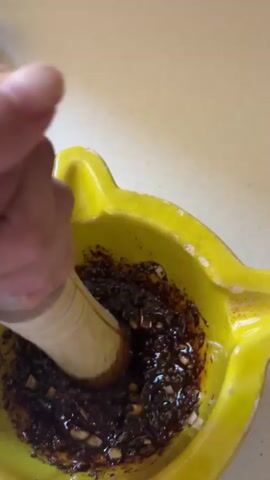 Here’s a detailed description of the video:

The video shows a close-up shot of a person using a wooden pestle to grind ingredients inside a yellow mortar. The mortar is a decorative piece with an unusual shape.  The ingredients in the mortar appear to be a mix of dark-colored solids and a liquid, possibly spices, herbs, and oil.  The person's hand is visible as they move the pestle in a circular motion, grinding the ingredients. The surface underneath the mortar looks like a kitchen counter or countertop. The camera angle is slightly above, looking down into the mortar as the ingredients are being mixed.