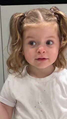 A young girl is seen in a close-up shot, dressed in a white t-shirt and with her blonde hair styled in pigtails. She looks directly at the camera, smiling slightly. An audio track of someone singing along with music plays in the background. The scene appears to be indoors, with a white or light gray wall behind her.