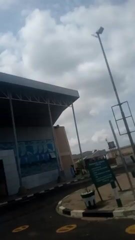 The video shows an outdoor scene with a building in the background that has a covered walkway. The building appears to be a commercial establishment, possibly an airport terminal or a large shop, with a decorative facade featuring blue and white patterns and the word "FAAN" visible on some panels. In the foreground, there are people, some wearing white shirts with green trim and green caps, suggesting a uniform. One person in the foreground is wearing a white shirt with "NSC" printed on the back and a green cap. Another person in the foreground is wearing a white shirt and a green cap. The ground is paved, and there are some yellow markings on the pavement. The sky is cloudy. The camera appears to be moving.