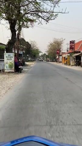 A first-person perspective video captures a person riding a motorcycle down a paved road lined with buildings and trees. The sky is overcast and bright. On the left side of the road, there is a pile of tires and a motorcycle parked next to a building. Further down the road, a few people can be seen standing near the parked motorcycle. On the right side of the road, there are various shops and buildings, some with signs. Several vehicles, including trucks and cars, are traveling in the same direction as the motorcycle. The road appears to be a rural or suburban area.