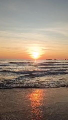 The video shows a beach at sunset. The sun is setting on the horizon, casting an orange glow over the sky and water. There are gentle waves coming onto the shore, and the wet sand reflects the sunlight. In the distance, a pier or some other structure can be seen. There are no people visible in the video.