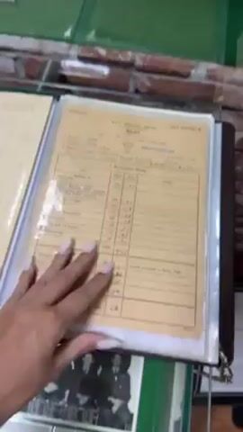A person's hand with long fingernails flips through documents inside a binder on a glass-covered surface. The documents are old and appear to be official records related to the Royal Air Force. The first visible document is a "Grading Report PILOT" form. The subsequent document is a "Royal Air Force NOTICE PAPER." The background includes a brick pattern and a green surface, suggesting the items are on display.