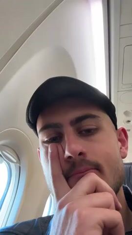 The video shows a man sitting in an airplane seat. He is wearing a black cap and a black leather jacket. He appears tired or pensive, resting his face on his hand. The camera then pans to the window of the plane, revealing a view of the tarmac on a clear day. We can see airport infrastructure such as buildings, vehicles, and light poles. The airplane is stationary on the ground.
