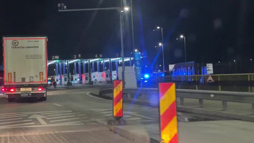 At night, a vehicle films from the driver’s seat, approaching toll booths. Two large trucks are ahead, one pulling into a toll booth. The toll booths are well-lit, and road signs are visible. The scene is dark, illuminated by the vehicle's headlights, the toll booth lights, and streetlights. The vehicle appears to be moving slowly.