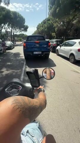 The video begins with a first-person perspective from a scooter, showing the handlebars and a tattooed arm. The rider is looking forward down a street lined with trees and parked cars. A blue Ford Ranger pickup truck is in front of the scooter, moving in the same direction. The license plate of the truck reads "0518 NFN". Several other cars are parked along the street. The sun is shining, casting shadows. The video progresses slightly, showing the truck continuing down the road and the scooter following. The environment appears to be a sunny, outdoor setting on a paved road.
