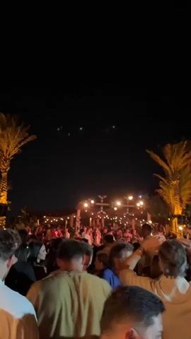 A nighttime aerial view shows a large gathering of people, many of whom appear to be enjoying themselves, possibly at an outdoor event or party. Tall palm trees are visible on the left and right sides of the frame, illuminated from below, casting a warm glow. String lights hang in the background, illuminating a building or structure. In the dark sky above, several small, bright green lights are visible, arranged in clusters and scattered patterns. The lights appear to be in motion, with some forming distinct shapes or lines. The overall atmosphere is festive and mysterious due to the unusual lights in the sky.
