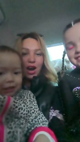 Three people, a woman and two young girls, are in a car. The woman is in the middle and appears to be recording a video. To her left is a toddler wearing a leopard print outfit with a pink trim. To her right is a young girl with braided hair, wearing a black outfit with colorful rhinestones, and pink makeup. The car interior is visible in the background. The lighting suggests it is daytime. The toddler is making sounds while the woman is speaking. The older girl is smiling.