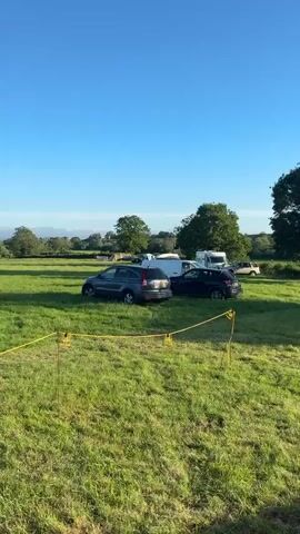 The video shows a grassy field under a clear blue sky with some trees and distant hills. Several vehicles are parked in the field. In the foreground, a yellow rope is stretched between two yellow posts. A dark grey SUV is parked to the left of the frame, and a dark colored SUV is parked to its right. Further back, a white van and what appears to be a camper van are visible. The camera pans slowly across the scene.