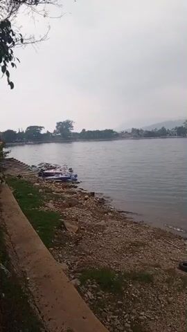 The video pans across a body of water with a rocky shore. On the shore, several small boats are docked. In the background, trees and buildings are visible, with hills or mountains in the distance under a cloudy sky. The camera moves from left to right, showing more of the water and the distant shoreline.