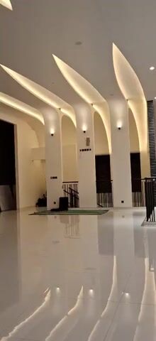 The video pans across the interior of a modern building with a high, curved ceiling featuring embedded warm lighting. White columns support the ceiling. The floor is highly polished and reflective. In the foreground on the left, a green prayer rug is laid out, with a person in dark clothing kneeling on it. A black railing runs along a mezzanine level. On the mezzanine, two individuals are visible. One is wearing a dark garment and standing, while another, a child, is walking away from the camera. A tall, textured pillar with a dark pattern is situated on the mezzanine.